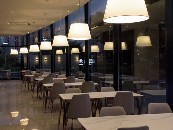 Un restaurant moderne avec une rangée de tables et de chaises, de grandes suspensions et une vue sur la ville grâce à de larges fenêtres.