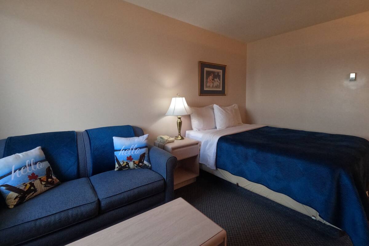 Habitación de hotel con un sofá azul, una mesita, una cama con una manta azul, una mesita de noche con una lámpara y un cuadro en la pared.
