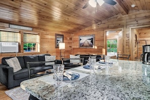 A cabin interior with a wooden ceiling, a black sofa, a dining table with plates and glasses, and a framed picture on the wall.