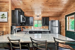 A kitchen with wooden walls, a granite countertop, black cabinets, stainless steel appliances, and a window with a view of greenery.