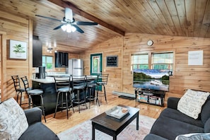 A cozy cabin-style room with a wooden interior, a dining area with bar stools, a television, and a view of a lake through the window.