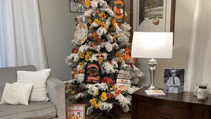 A living room with a decorated Christmas tree, a grey sofa, a wooden coffee table, a lamp, and a framed photo on the wall.