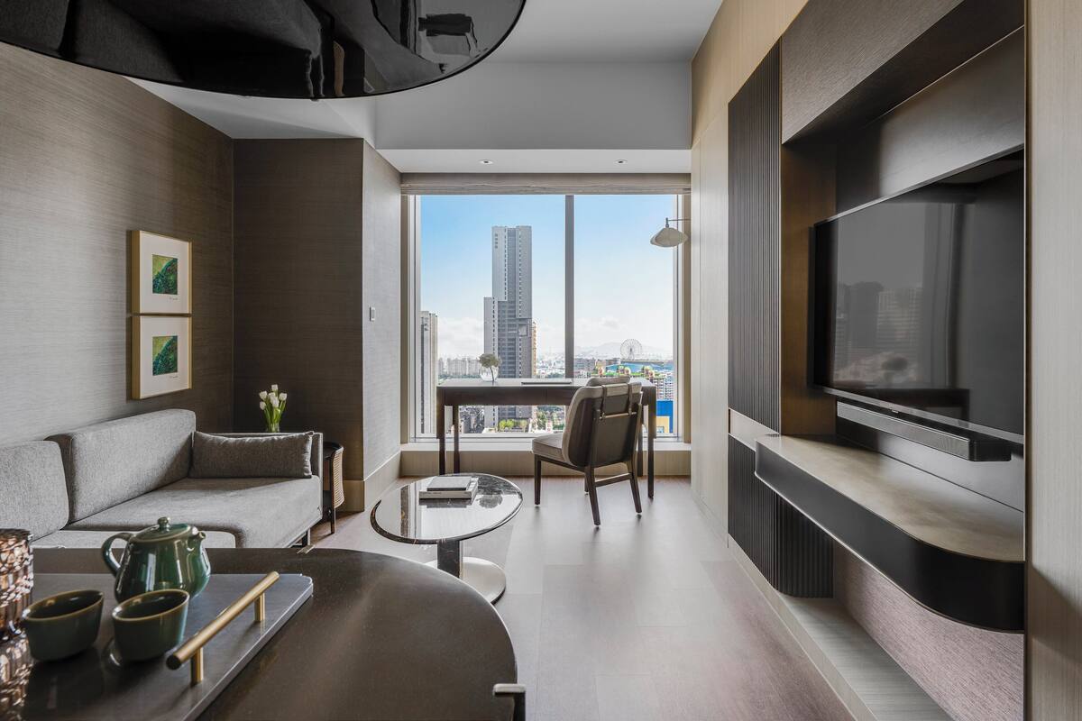 Ein modernes Wohnzimmer mit einer Couch, einem Couchtisch und einem Fernseher. Es verfügt über ein großes Fenster mit Blick auf die Stadt.