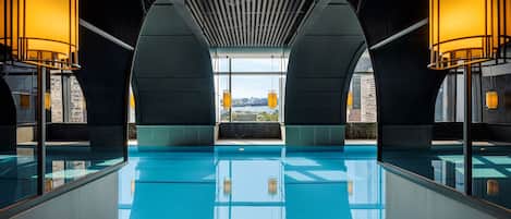 A modern indoor swimming pool with clear blue water, large windows, and warm lighting.