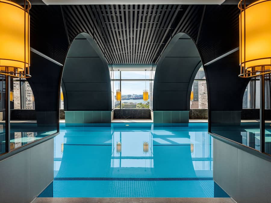 A modern indoor swimming pool with clear blue water, large windows, and warm lighting.