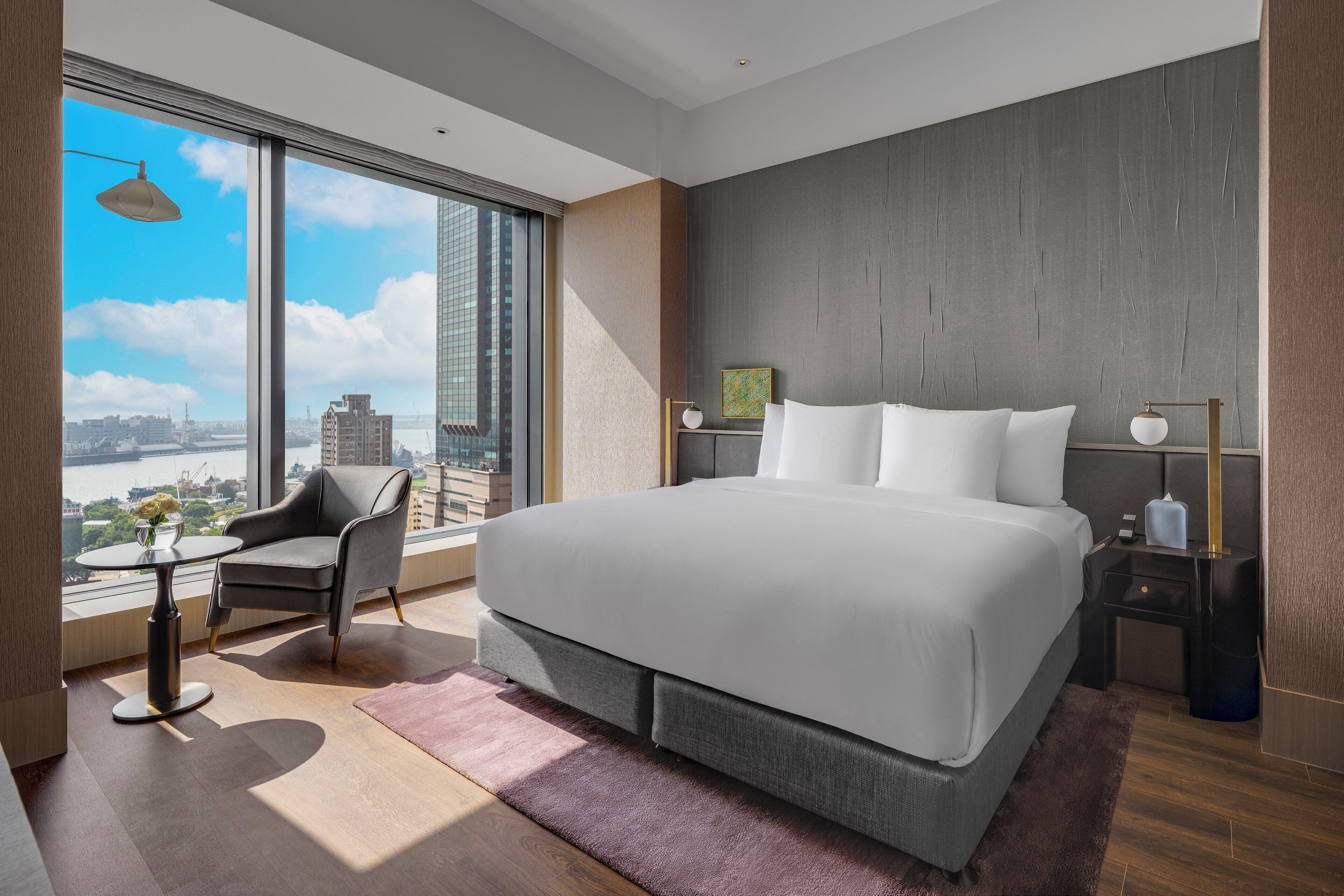 Una habitación de hotel moderna con una cama grande, una zona de estar con una silla, una mesita y vistas a la ciudad a través de ventanas amplias.