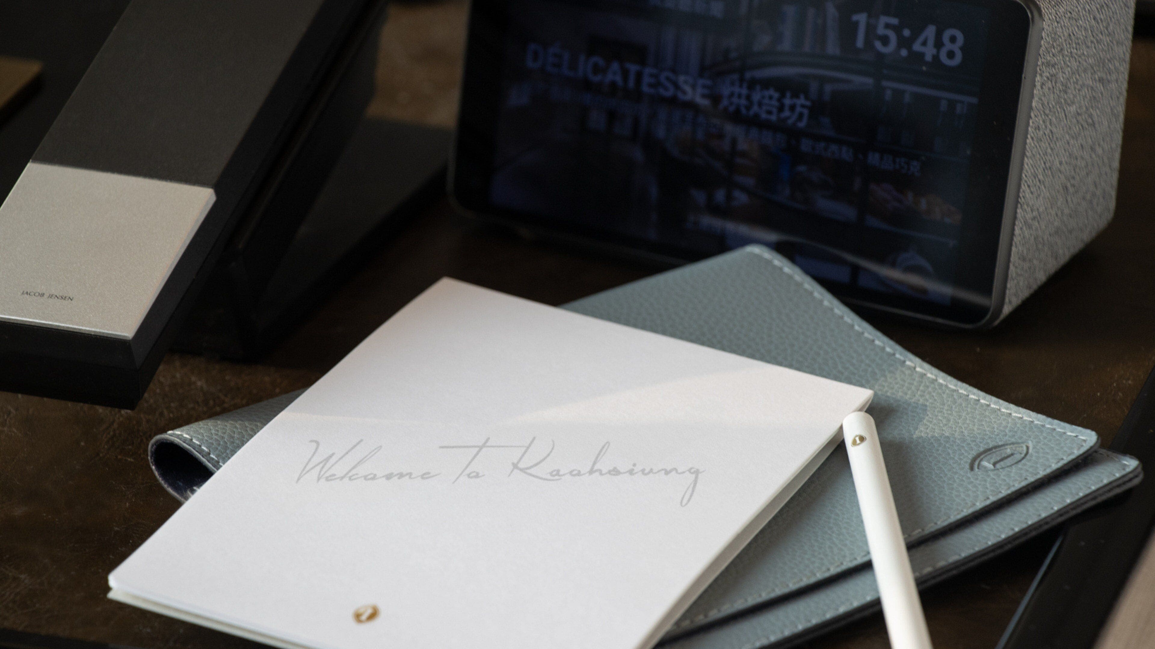 A notebook with a handwritten note, a pen, a digital clock, and a black box with 'JACOB JENSEN' on it.