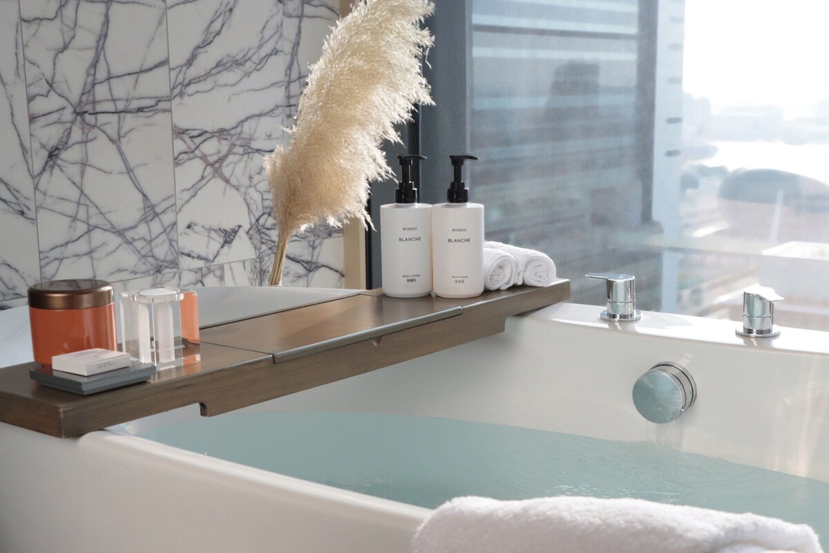 A modern bathroom with a marble wall, a bathtub with a view, and toiletries on a shelf.