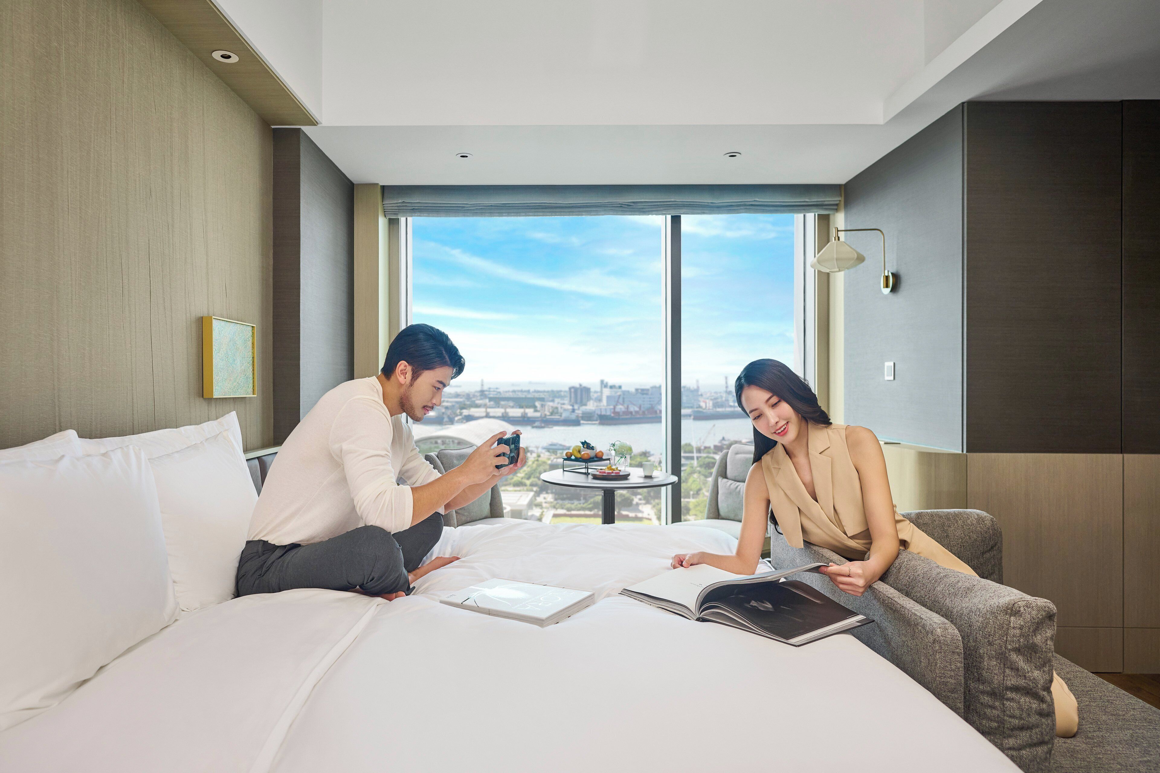 Une chambre d’hôtel moderne avec un grand lit, une petite table sur laquelle se trouve un panier de fruits, et une vue sur le paysage urbain à travers de grandes fenêtres.