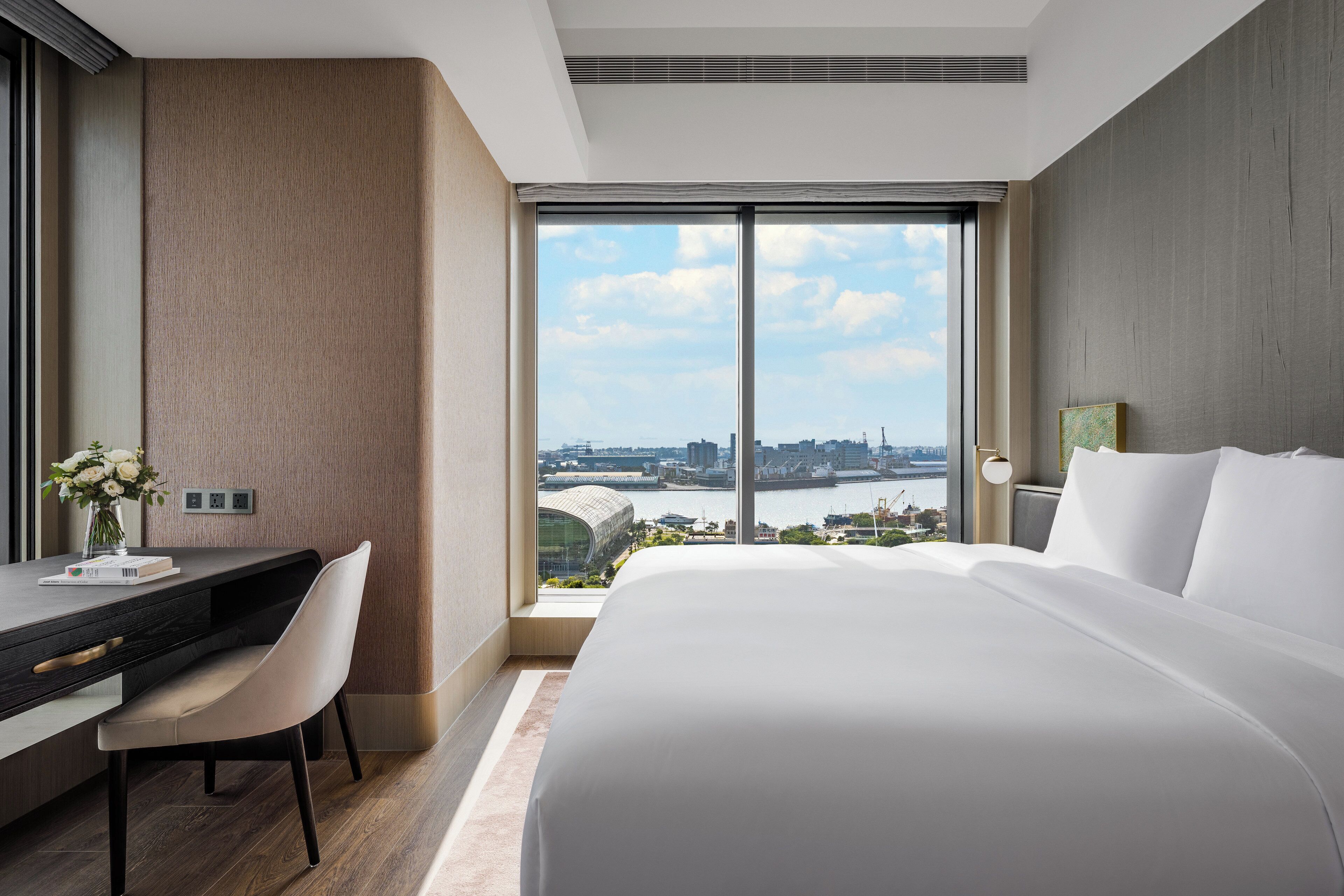 A hotel room with a large bed, a desk, and a chair. There is a view of a cityscape through the window.