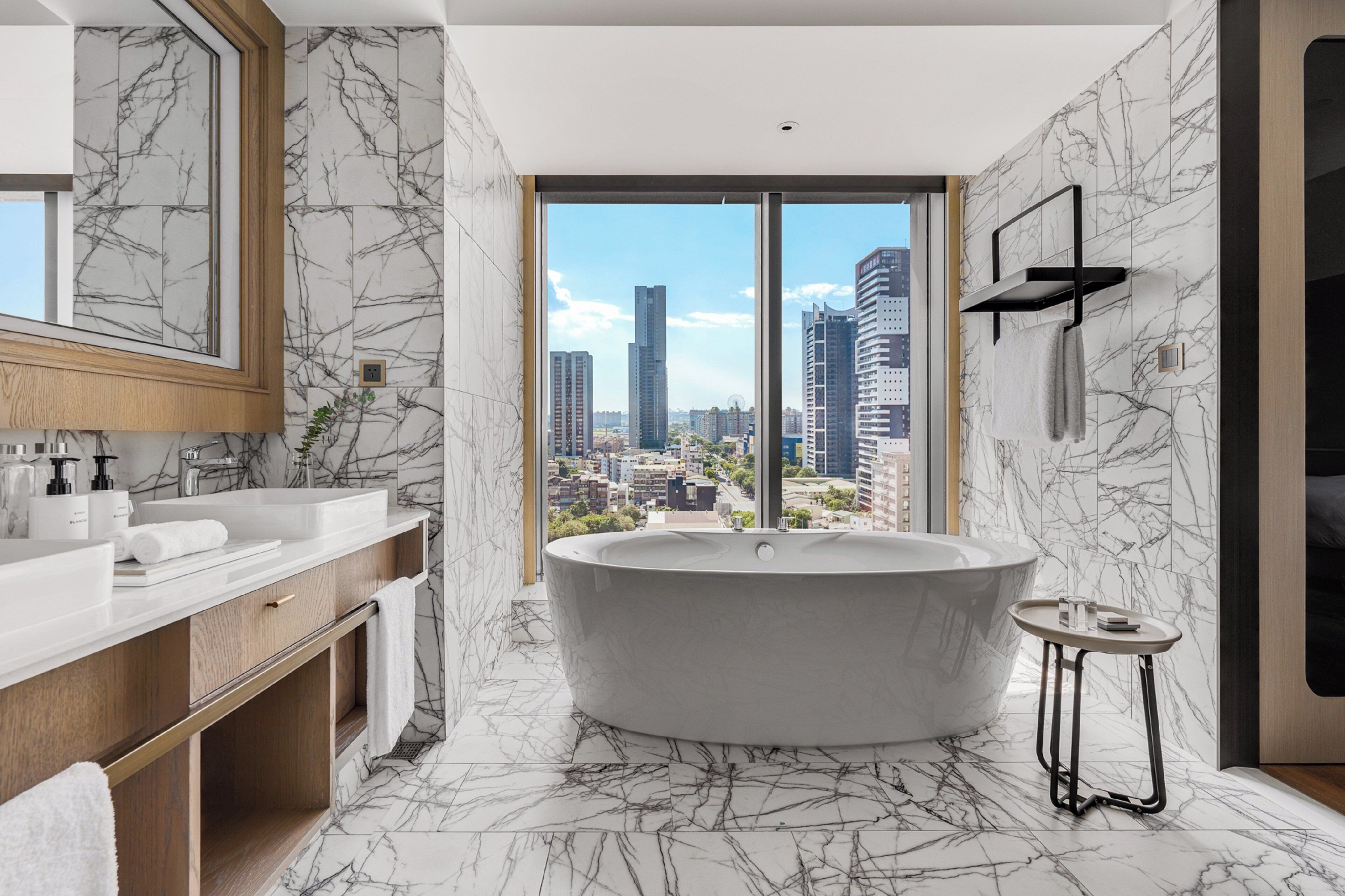 A modern bathroom with a freestanding bathtub, a large mirror, and a city view through floor-to-ceiling windows.