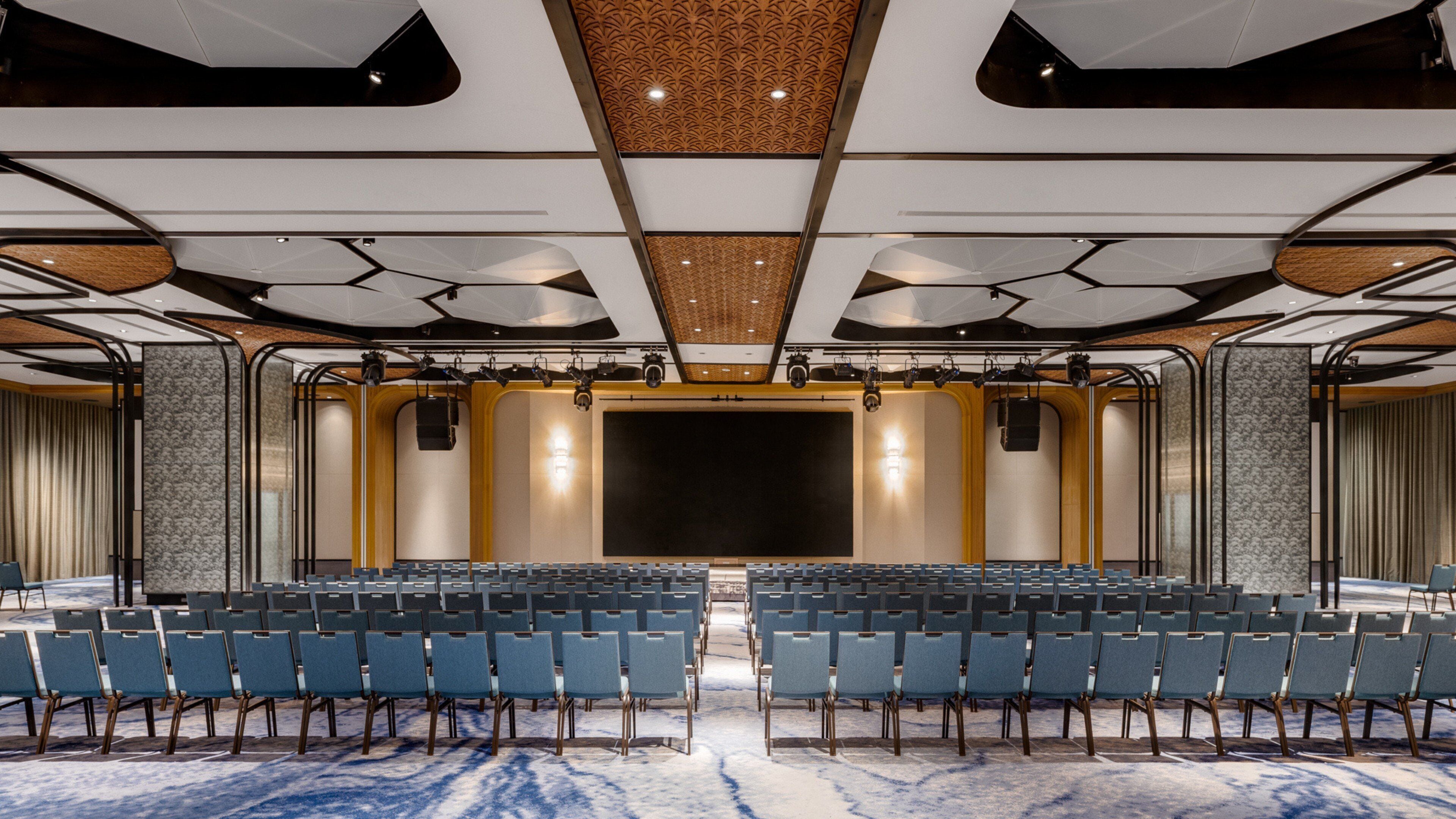 Een vergaderzaal met rijen stoelen, een podium en een versierd plafond.