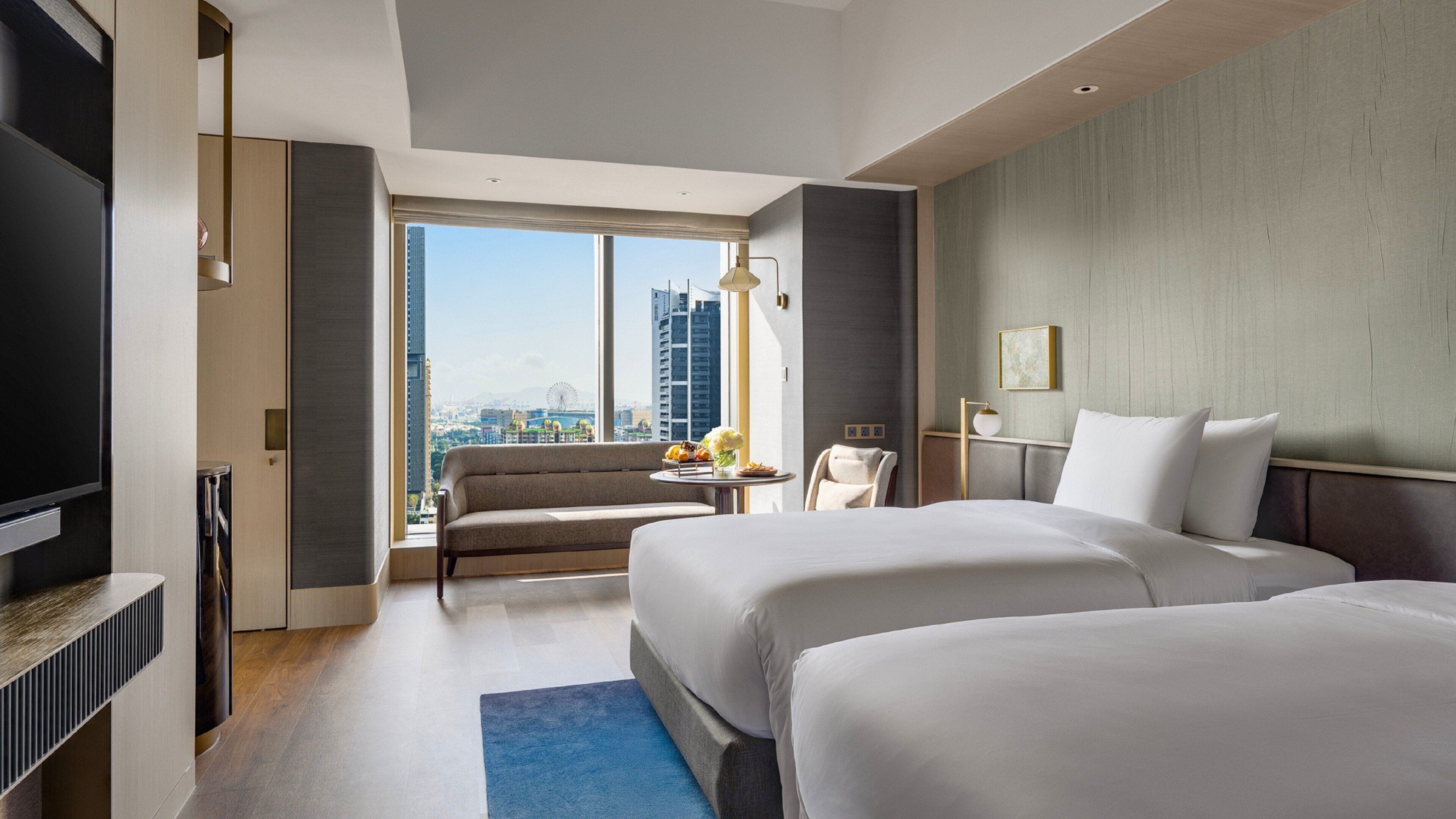 A hotel room with two beds, a sofa, a TV, and a view of the cityscape.