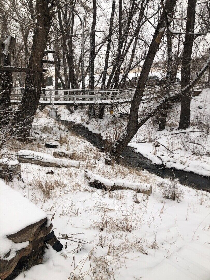 A snow-covered landscape with a wooden bridge over a stream, surrounded by trees and a stone wall.