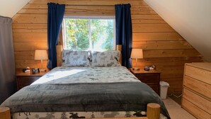 A cozy bedroom with a sloped ceiling, a bed with a moose-themed duvet, two bedside lamps, a wooden nightstand, and a dresser. - Moose Tracks Cabin in North Pole, Alaska (North Pole)