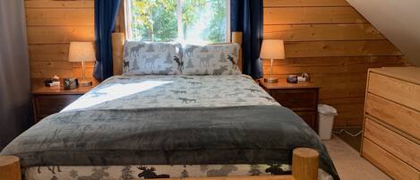 Une chambre confortable avec un plafond en pente, un lit avec une couette à motifs d’élan, deux lampes de chevet, une table de chevet en bois et une commode.