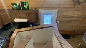 A wooden cabin interior with a kitchen area, a small refrigerator, and a window with a view of trees. - Moose Tracks Cabin in North Pole, Alaska (North Pole)