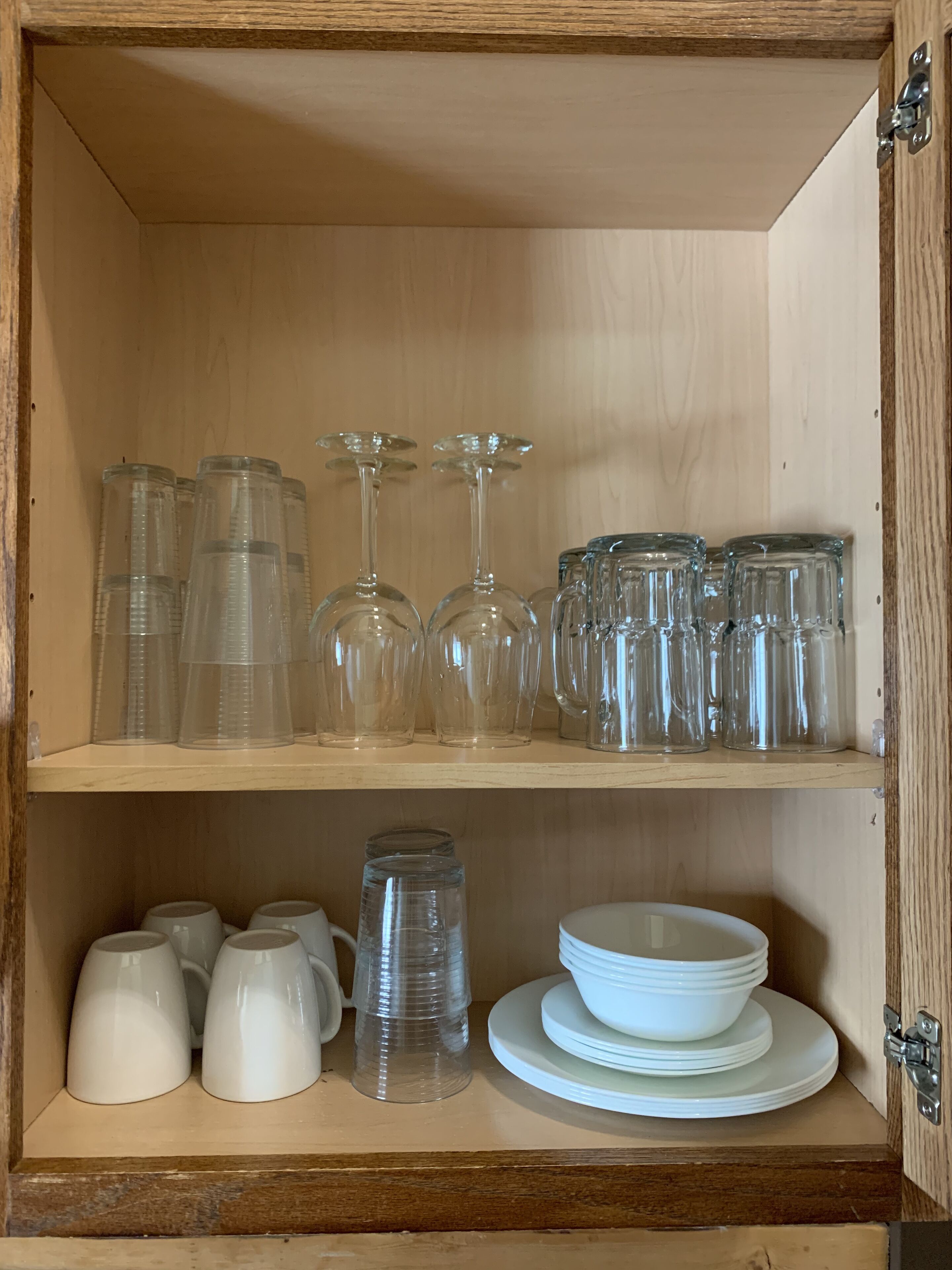 Un mueble de madera con estantes que contienen vasos de plástico transparente, copas de vino y vajilla de cerámica blanca.