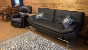 A wooden interior with a black leather sofa, two matching armchairs, a patterned rug, and a window with curtains. - Moose Tracks Cabin in North Pole, Alaska (North Pole)