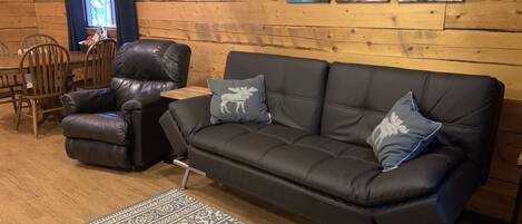 Un intérieur en bois avec un canapé en cuir noir, deux fauteuils assortis, un tapis à motifs et une fenêtre avec des rideaux.