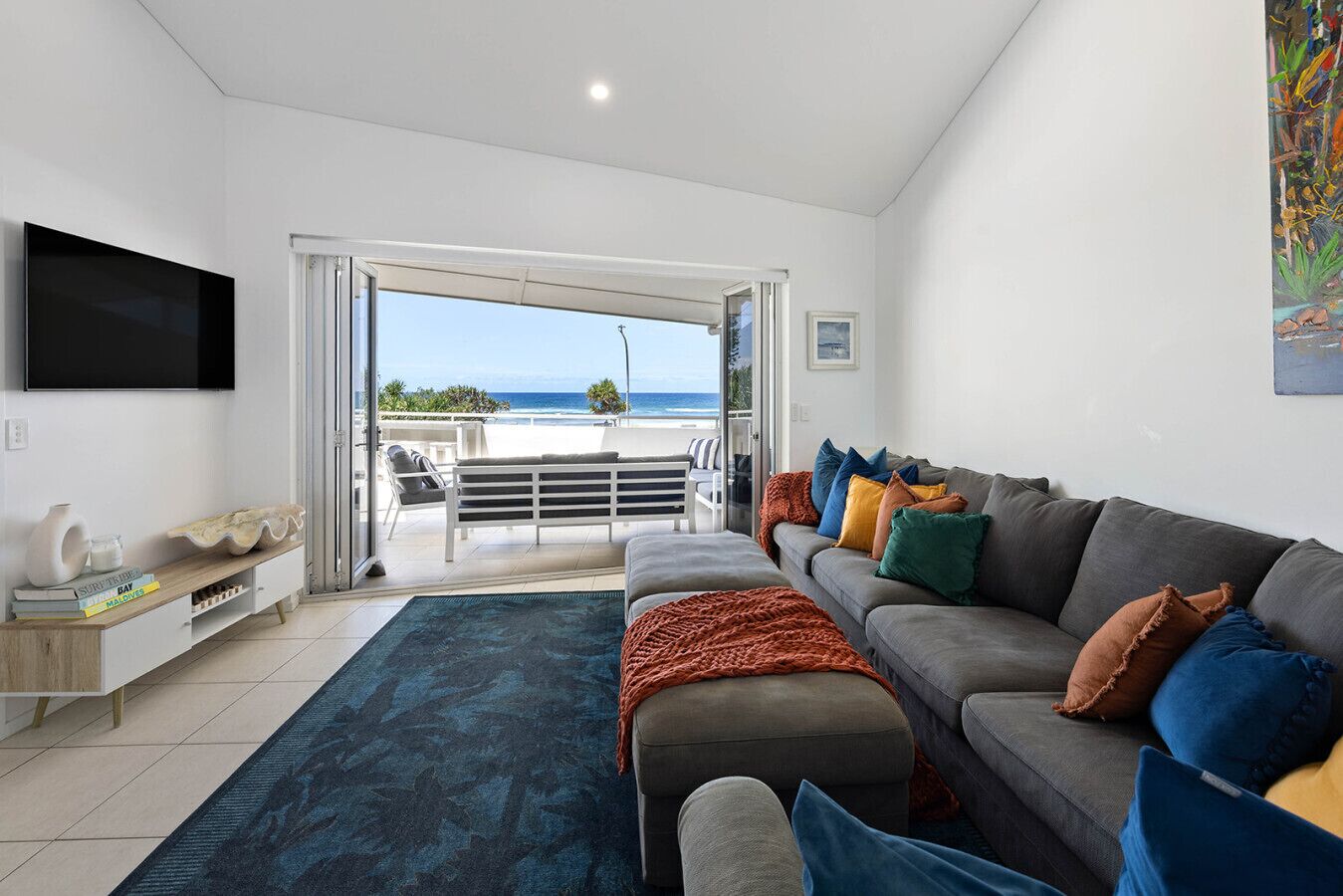 A modern living room with a grey sofa, colorful cushions, a flat-screen TV, a wooden console, and a view of the ocean through a sliding glass door.