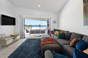 A modern living room with a grey sofa, colorful cushions, a flat-screen TV, a wooden console, and a view of the ocean through a sliding glass door. - Lennox Head Waves Beachfront Penthouse (Lennox Head)