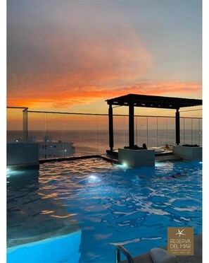 Una piscina infinita con vista al atardecer, un área de estar cubierta y una barandilla de cristal.