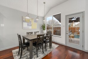 A dining area with a wooden table and chairs, a large window, and a view of a backyard with a pool. - Fancy home in Frisco-Heated spa &pool BBQ Gameroom close to DFW sleep 10-14 (Frisco)