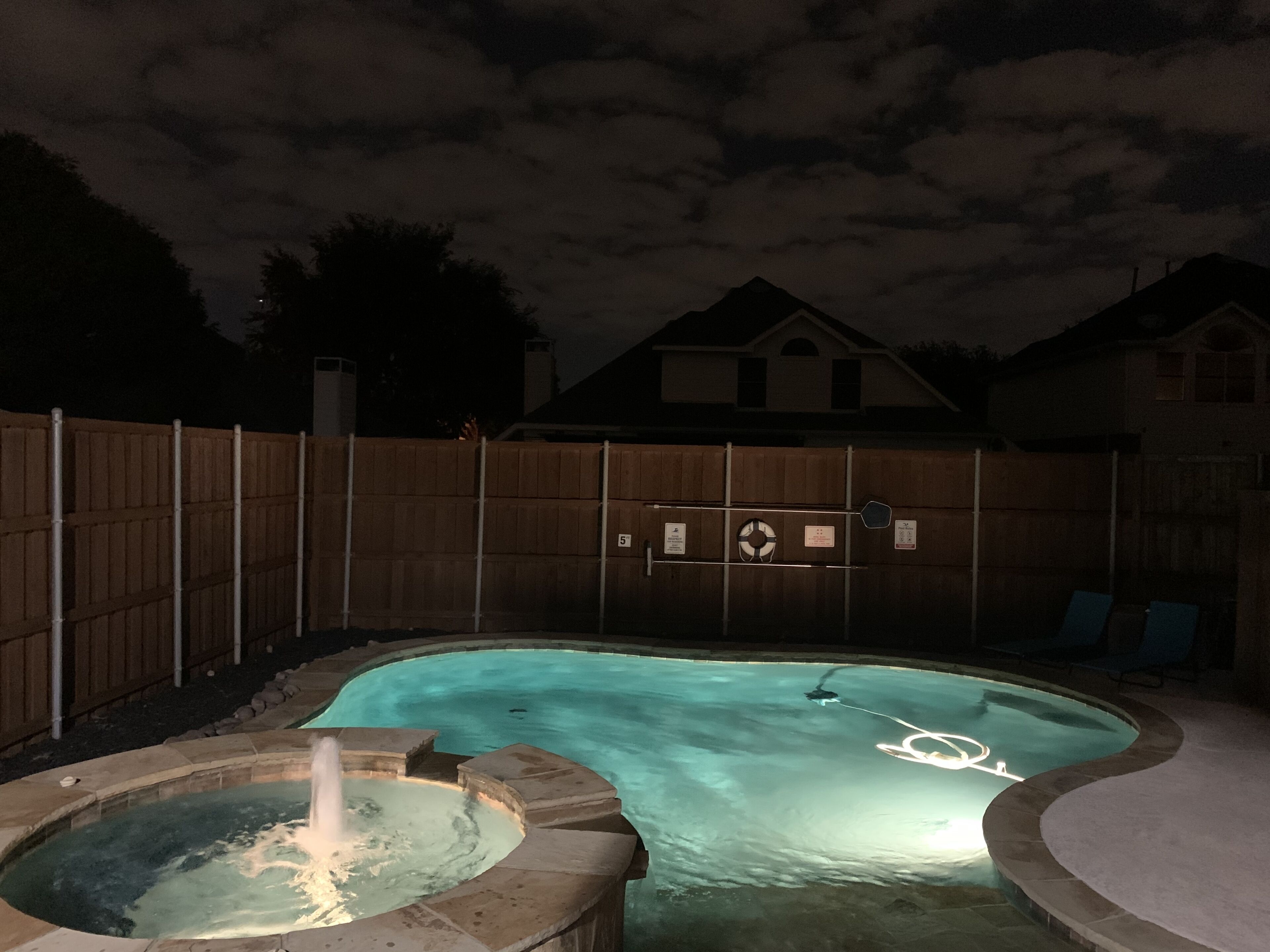 A backyard swimming pool with a hot tub, surrounded by a wooden fence, and a house in the background.