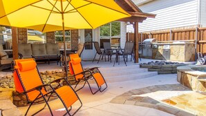 Une terrasse extérieure avec un sol en pierre, un barbecue et des chaises orange sous un parasol jaune.