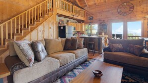 A spacious living area with a wooden staircase, a kitchen in the background, and a large window providing natural light. - Comfy Lodge with Stunning Mountain Views (Waynesville)