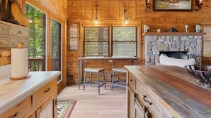 A wooden cabin interior with a stone fireplace, wooden bar counter, and two bar stools. - Comfy Lodge with Stunning Mountain Views (Waynesville)