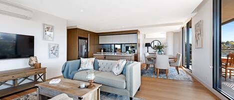A modern living room with a sofa, coffee table, and TV. There is a kitchen in the background with a dining area.