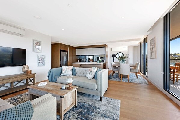 A modern living room with a sofa, coffee table, and TV. There is a kitchen in the background with a dining area.
