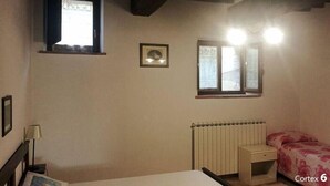 A small, sparsely furnished room with a bed, a desk, a radiator, and two windows. - Apartment Ortensia 1 Bedroom, Living Room,bathroom (Monte Castello di Vibio)