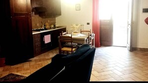 A living room with a dark-colored sofa, a dining table with chairs, a kitchen area with cabinets and a stove, and a bright doorway leading outside. - Apartment Ortensia 1 Bedroom, Living Room,bathroom (Monte Castello di Vibio)