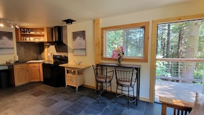 A kitchen with a stove, sink, and wooden cabinets. There is a window with a view of trees and a door leading to an outdoor area. - Beautiful cottage, close to beach (Port Orford)