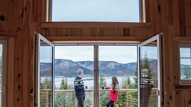 Una cabaña de madera con un amplio ventanal corredera que da a una terraza con vista a montañas y un lago.