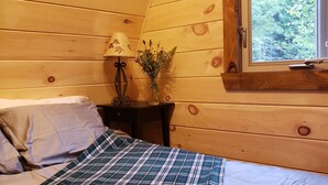 Un dormitorio acogedor con techo de madera, ventana con vistas a árboles, mesita de noche con lámpara y un jarrón de flores, y una cama con manta escocesa.