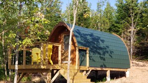 Una cabaña de madera con techo curvo, una terraza con sillas y un sendero de tierra que lleva a ella.