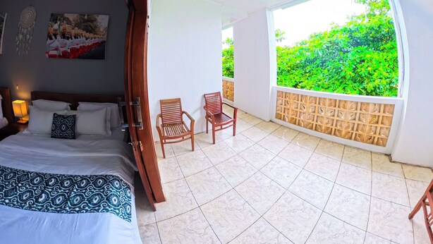 A bedroom with a bed, a nightstand, and a chair. There is a balcony with a view of greenery. - Ketapang Guesthouse Echo Beach Canggu (Canggu)
