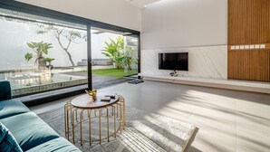 A modern living room with a large glass door leading to a garden, a round gold table, a blue sofa, and a flat-screen TV mounted on the wall. - Mile Stone Villa (Jimbaran)