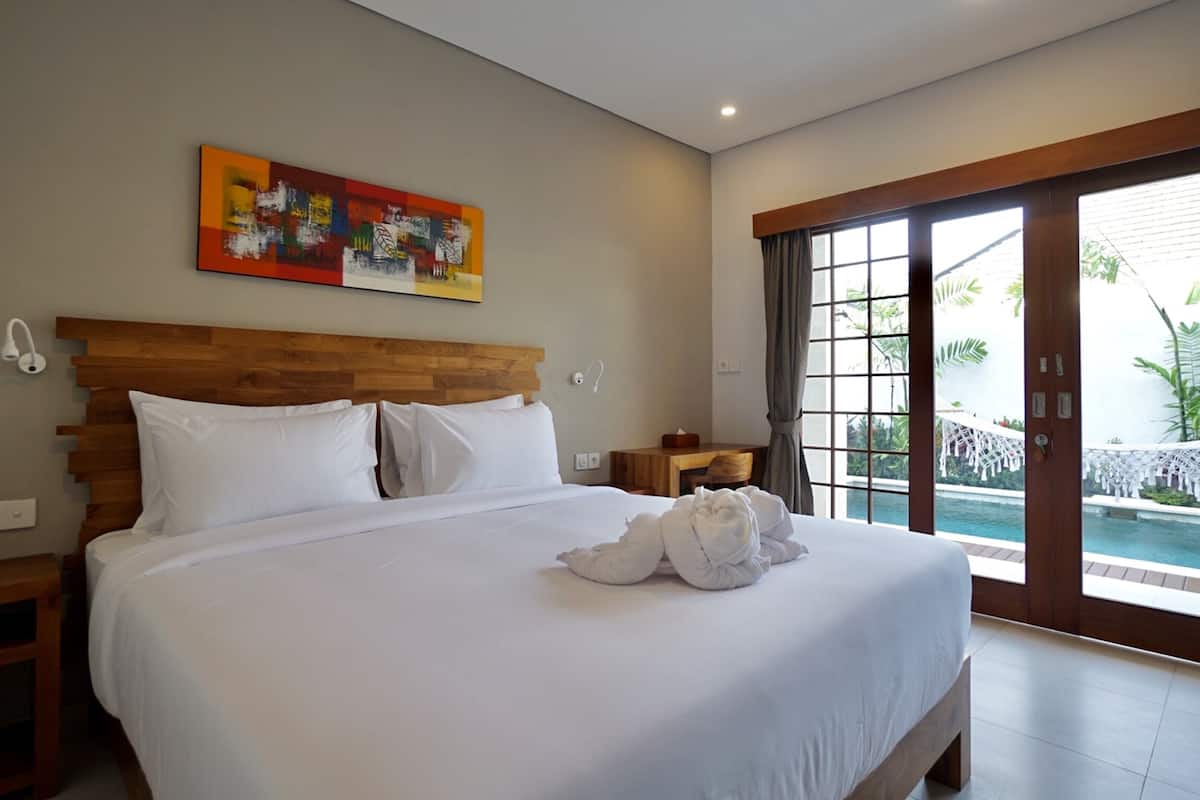 A large bed with white linens, a wooden headboard, and a painting above it. There is a window with a view of greenery and a pool.