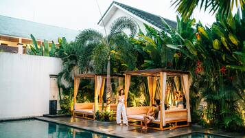 Un área junto a la piscina con un espacio de descanso cubierto por una pérgola, piscina y exuberante vegetación tropical.