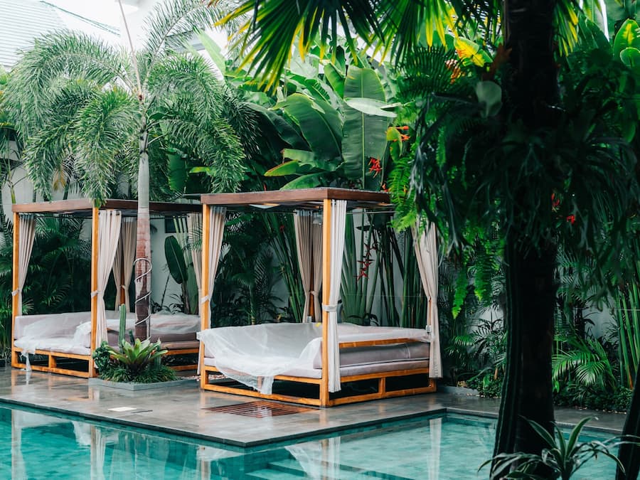 Deux lits à baldaquin en bois, placés au bord d’une piscine et entourés d’une végétation tropicale luxuriante.