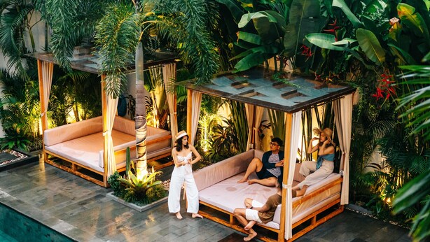 A poolside area with two wooden cabanas, one with a white and one with a brown cushion, surrounded by lush tropical plants. - The Kamare by Zuzu (Tibubeneng)