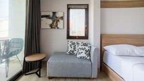 A modern hotel room with a grey sofa, a small round table, a wooden headboard bed, and a large window with blinds. - Little Nuts Villa (Jimbaran)