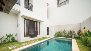 A swimming pool with a tiled deck, surrounded by a white building with a sliding glass door and a small patio area with plants. - Little Nuts Villa (Jimbaran)