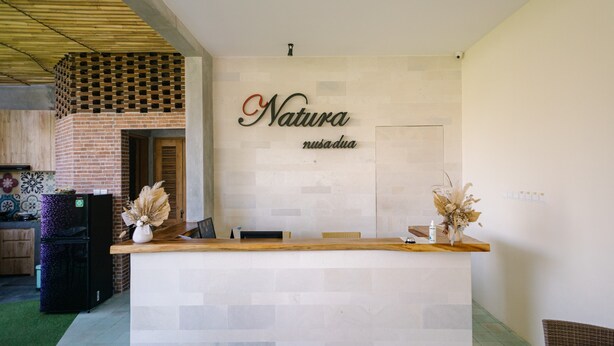 A reception desk with a sign that reads "Natura nusadua," a brick oven, and a kitchen area with a refrigerator. - Natura Nusa Dua (Nusa Dua)