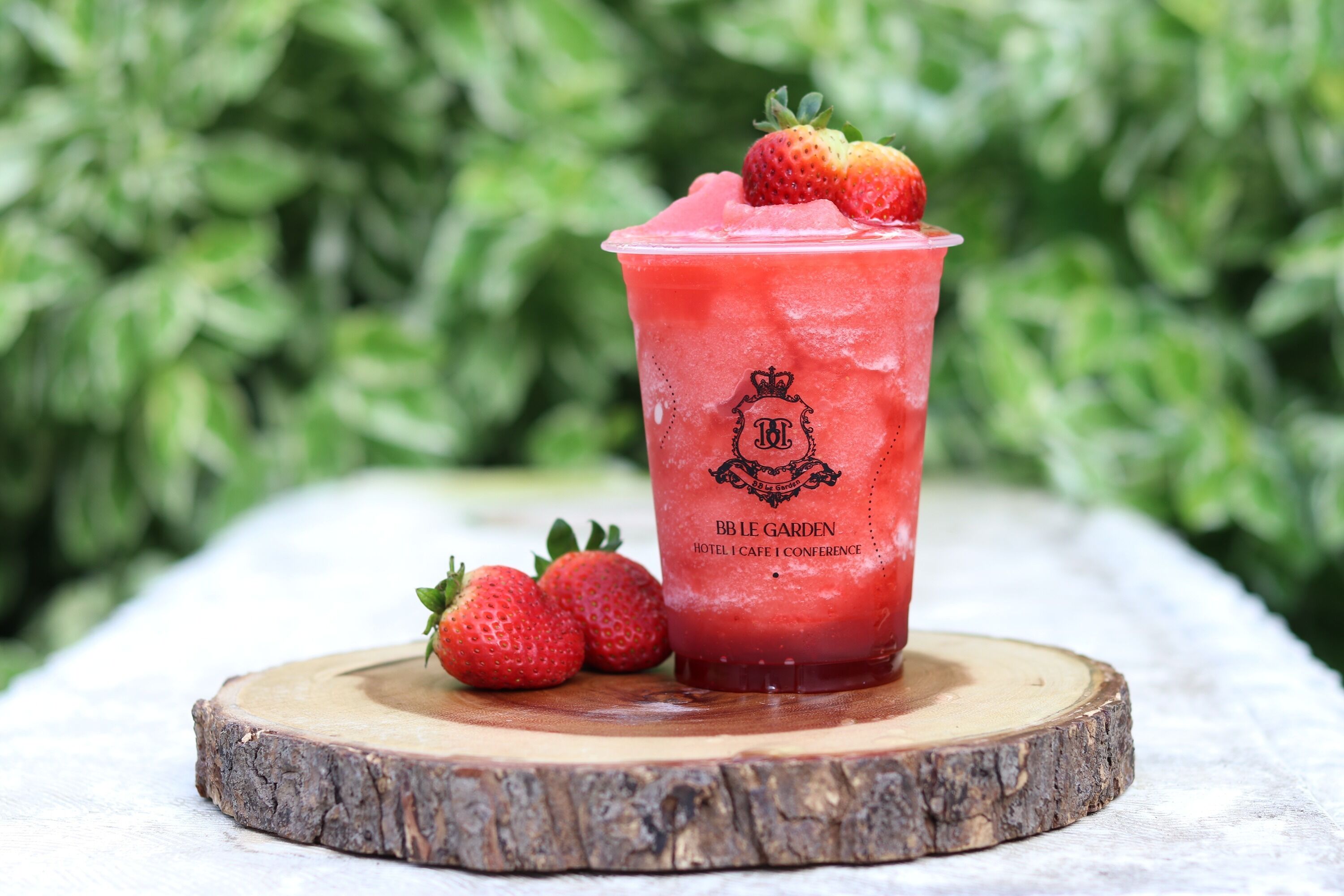 A glass of strawberry smoothie on a wooden coaster with a logo, placed on a white surface with green foliage in the background.
